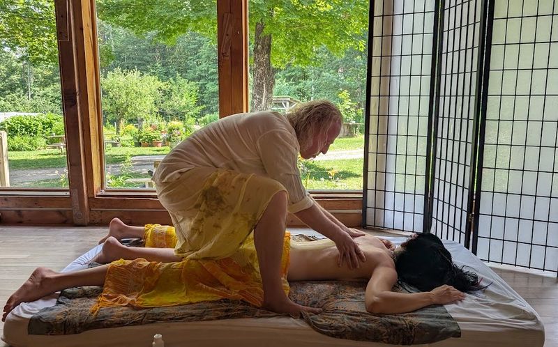 Sergio kneeling over a client who lies face down on a mattress by the windows, pressing along her back with both hands
