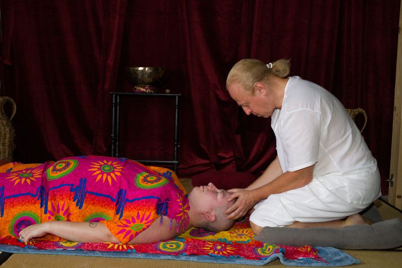 Sergio kneeling at the head of a client who lies on their back under a bright sarong, holding the client's head in his hands